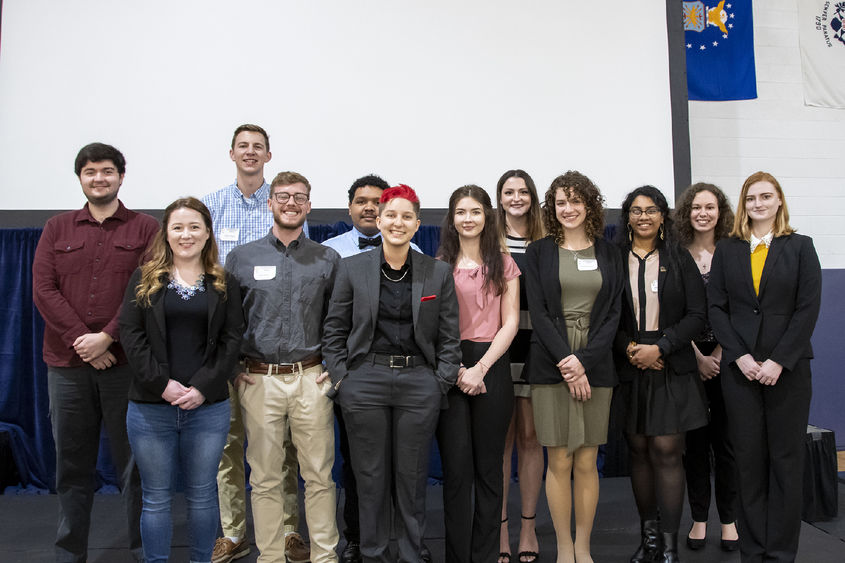 A dozen Penn State students pose for a photo on stage in front of a projector screen