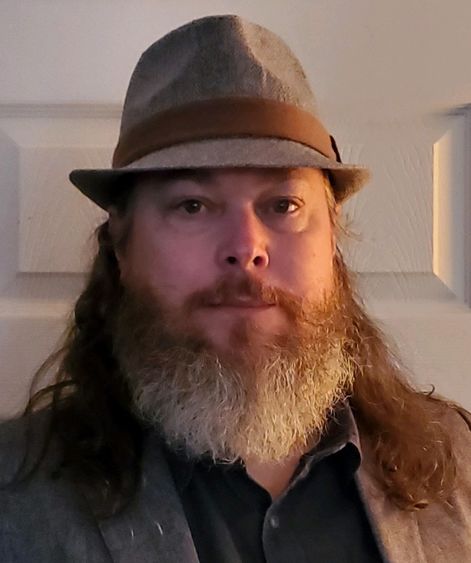 Man with a grey beard wearing tan fedora-style hat.