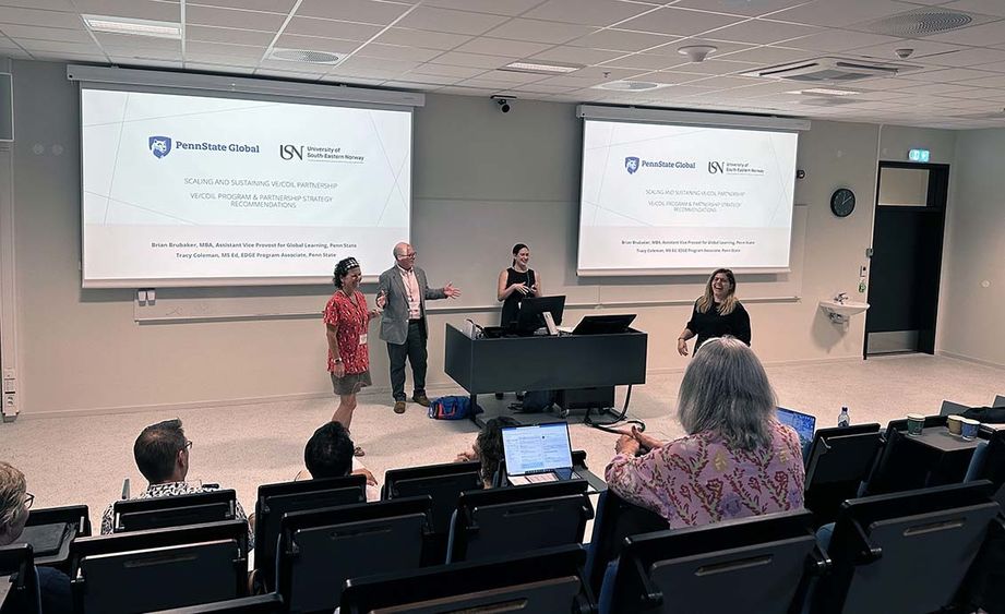 Four people standing in front of an audience in a classroom with two video projector screens on either side of them.