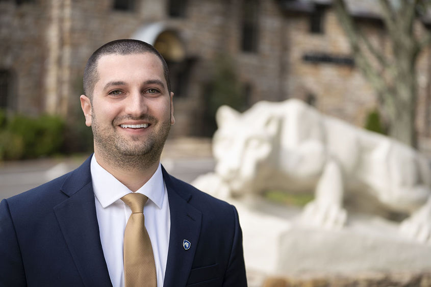 Man in a suit and tie smiling in front of Nittany Lion statue.