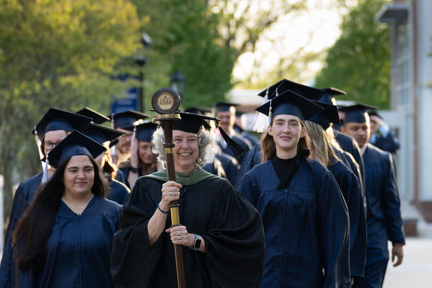 Graduates in caps and gowns in two rows walking down sidewalk led by faculty member in black robe carrying a staff and pendant.