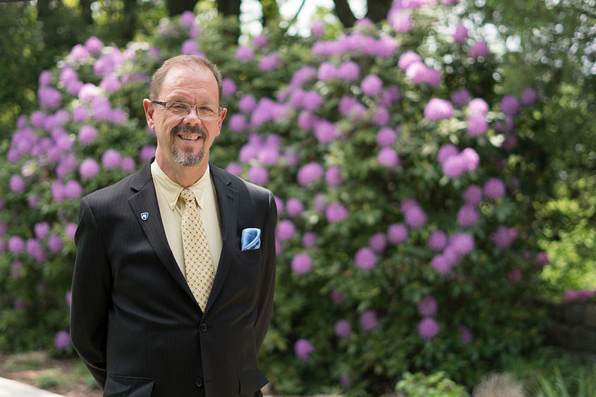 Man in a suit standing in front of a bush with purple flowers.