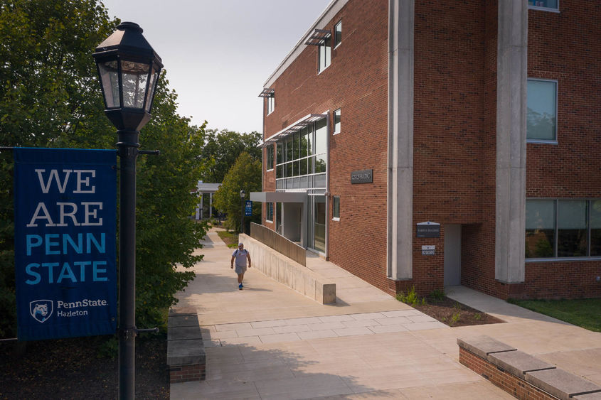 Student walking along sidewalk in between trees and a large brick building.