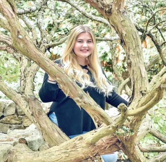 Blonde woman in a dark sweatshirt posing from behind tree branches.