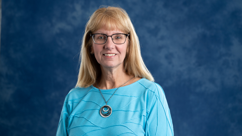 Woman with long blonde hair, glasses and light blue long-sleeved shirt.