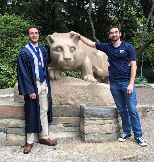 Male graduate in cap and gown and another male student standing next to Nittany Lion statue.